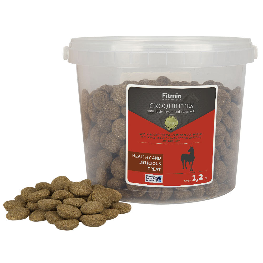 A 1.2kg transparent plastic bucket filled with Fitmin Horse Croquettes, with a small pile of the disc-shaped treats displayed in front. The red label highlights the apple flavour and Vitamin C content, designed as a healthy and delicious reward for horses.