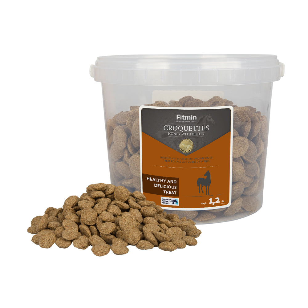 A 1.2kg transparent plastic bucket of Fitmin Horse Croquettes with honey and biotin, with a large pile of disc-shaped treats in the foreground. The orange and black label describes them as a healthy, easily digestible, and delicious treat for all horse categories.